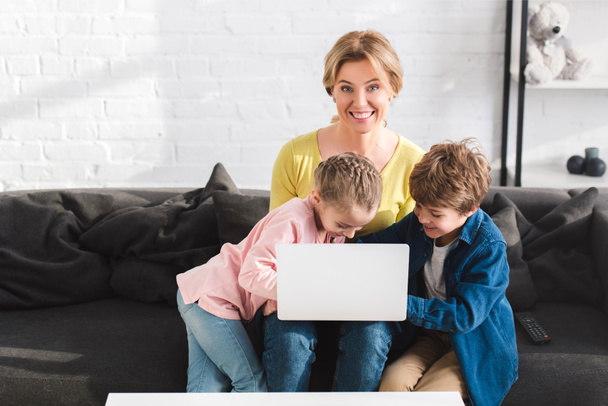 feliz madre sonriendo a la cámara mientras usa el ordenador portátil con los niños en casa
 - Foto, imagen