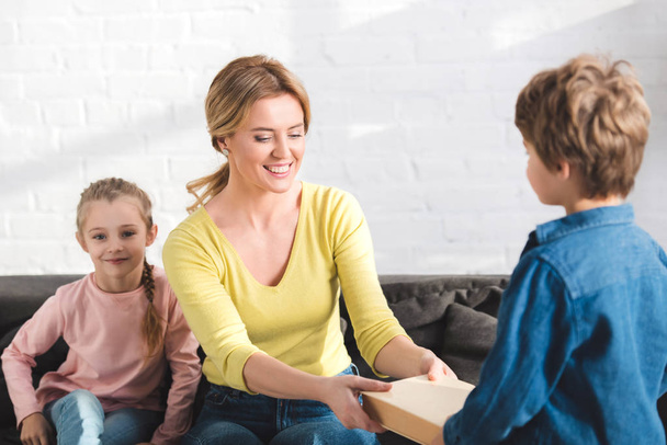 boy giving book to happy mother sitting on couch - Photo, Image