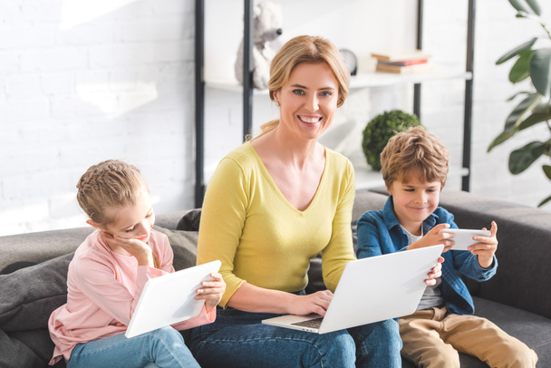 happy mother using laptop and smiling at camera while kids using digital devices at home - Foto, Imagem