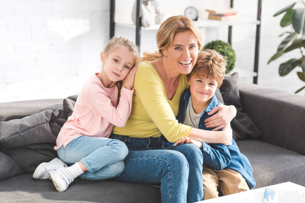 šťastný matka a rozkošné malé děti sedí na pohovce a usmívá se na kameru - Fotografie, Obrázek