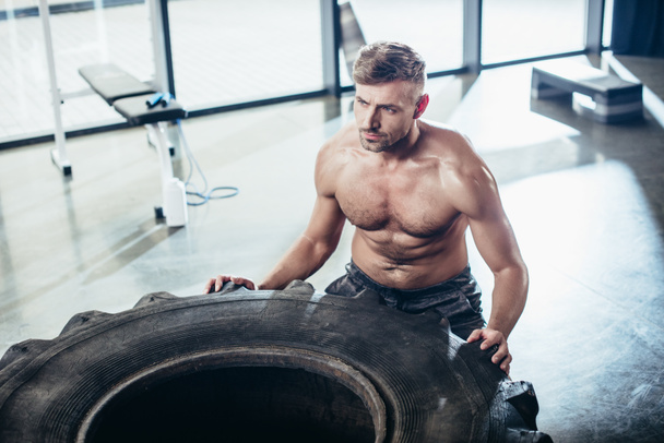 high angle view of handsome shirtless sportsman lifting tire in gym  - Fotoğraf, Görsel