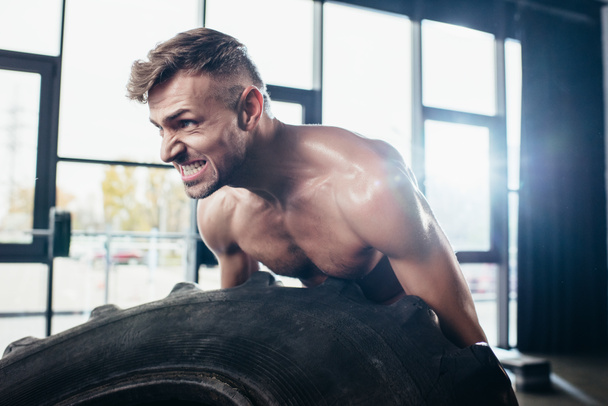 handsome muscular sportsman lifting tire and grimacing in gym  - Photo, Image