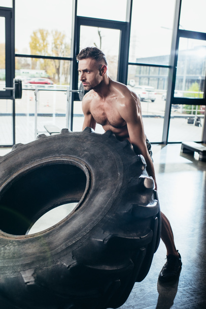 handsome shirtless sweaty sportsman lifting tire in gym  - Photo, Image