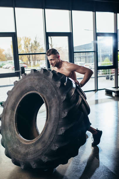 handsome shirtless sportsman lifting tire in sport center - Photo, Image