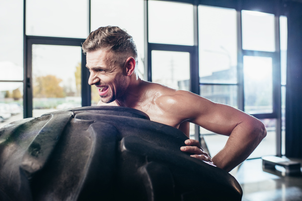 side view of handsome shirtless sportsman lifting tire and grimacing in gym  - Фото, зображення