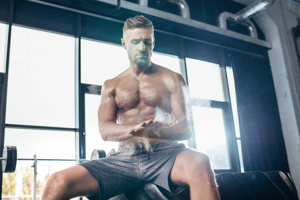 handsome shirtless sportsman sitting on tire and applying talcum powder on hands in gym  - Photo, Image
