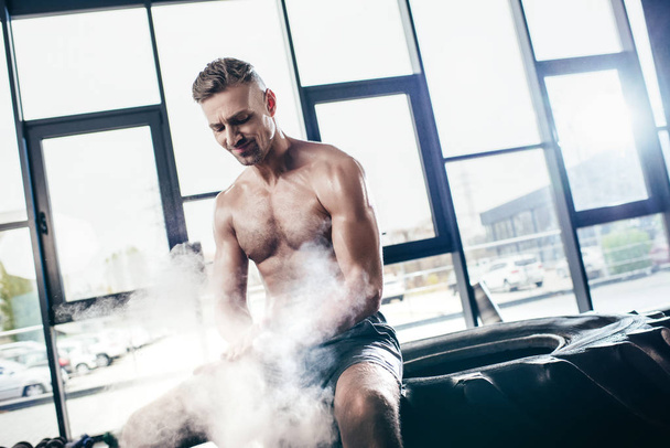 handsome cheerful shirtless sportsman sitting on tire and applying talcum powder on hands in gym  - Photo, Image