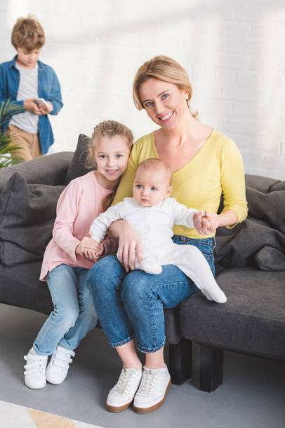 ευτυχισμένη μητέρα με βρέφος παιδί και αξιολάτρευτη κόρη κάθεται στον καναπέ και να χαμογελά στη φωτογραφική μηχανή - Φωτογραφία, εικόνα