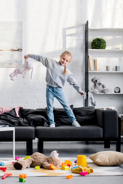 happy child standing on sofa and smiling at camera, various toys on floor - Fotografie, Obrázek