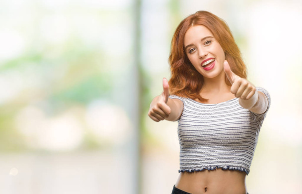Young beautiful woman over isolated background approving doing positive gesture with hand, thumbs up smiling and happy for success. Looking at the camera, winner gesture. - 写真・画像