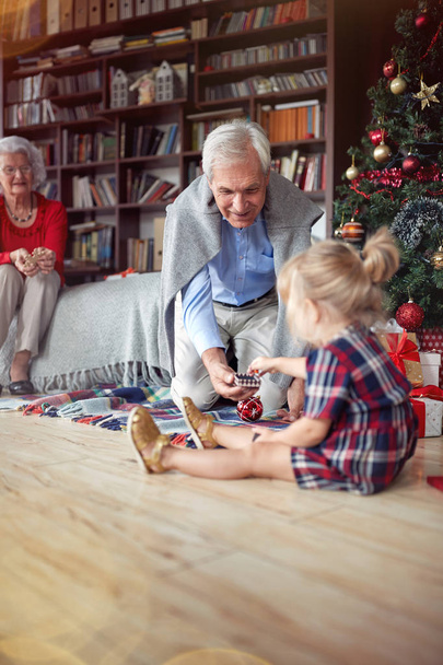 Cute girl is open Christmas present in front of a decorated X-mas tree with grandpa - Фото, изображение