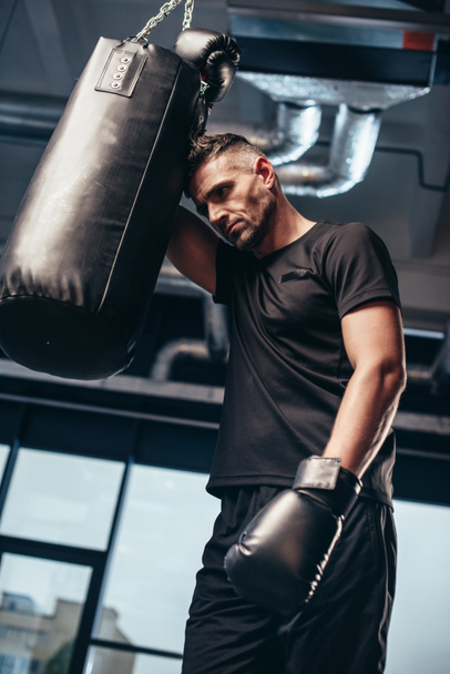 low angle view of handsome tired boxer leaning on punching bag in gym - Foto, afbeelding