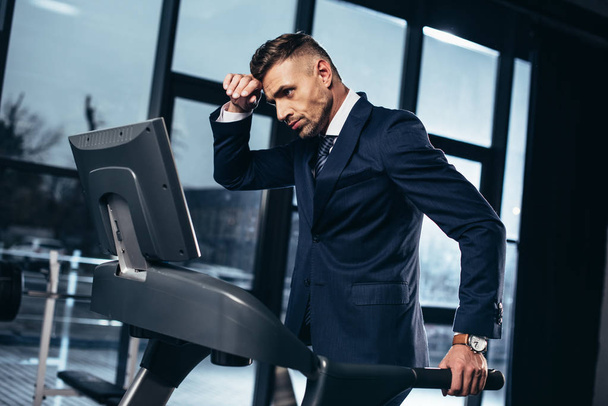 side view of tired handsome businessman in suit exercising on treadmill in gym - Fotoğraf, Görsel