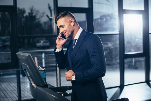 handsome businessman in suit exercising on treadmill and talking by smartphone in gym - Photo, Image