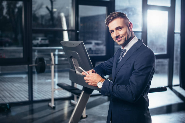 handsome businessman in suit leaning on treadmill and using smartphone in gym - Fotoğraf, Görsel