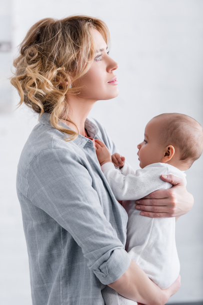 side view of thoughtful mother holding adorable infant child and looking away at home - Foto, immagini