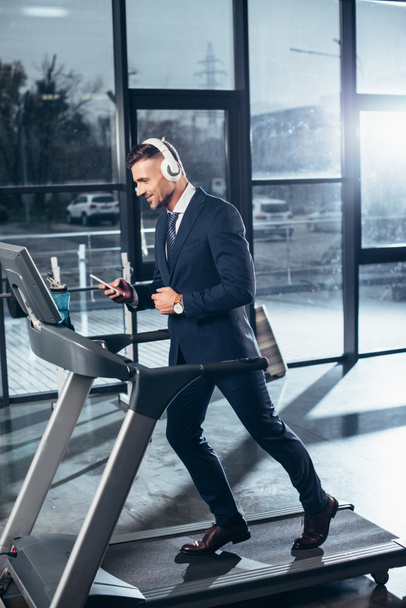 boční pohled pohledný podnikatel v obleku cvičení na běžeckém pásu a poslech hudby s smartphone v tělocvičně - Fotografie, Obrázek