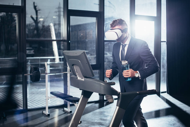 podnikatel v obleku a soupravu pro virtuální realitu cvičení na běžeckém pásu a drží láhev sport v tělocvičně - Fotografie, Obrázek
