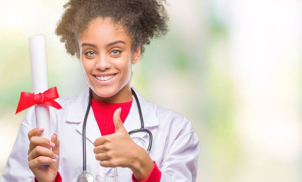 Young afro american doctor woman holding degree over isolated background happy with big smile doing ok sign, thumb up with fingers, excellent sign - Foto, Bild