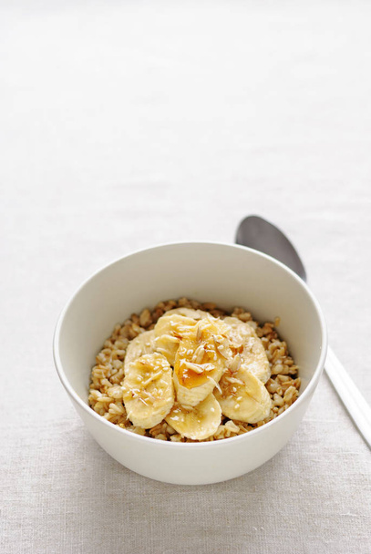 White ceramic bowl of oatmeal with banana slices and honey, served with the spoon on white table background. Viewed from high angle in closeup - Photo, Image
