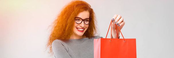 Portrait of beautiful redhead happy smiling young woman in gray clothes in glasses offering shopping bag to a client on gray background - Photo, Image
