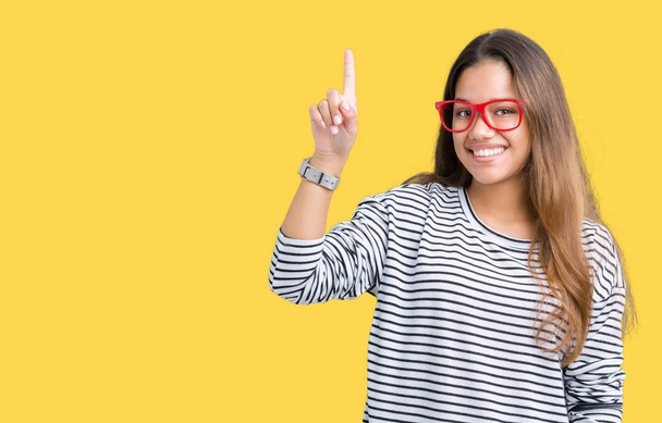 Young beautiful brunette woman wearing stripes sweater and red glasses over isolated background pointing finger up with successful idea. Exited and happy. Number one. - Photo, Image