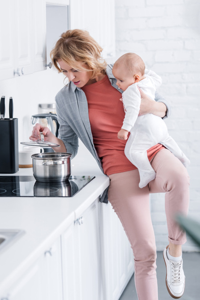 exhausted mother holding adorable infant child and cooking in kitchen - Valokuva, kuva