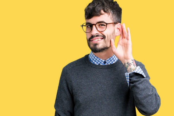 Mladý pohledný inteligentní muž nosí brýle izolované pozadí s úsměvem s rukou přes ucho naslouchá slyšení zvěsti nebo drby. Hluchota koncept. - Fotografie, Obrázek