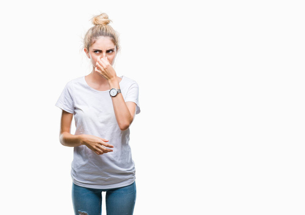 Young beautiful blonde woman wearing white t-shirt over isolated background smelling something stinky and disgusting, intolerable smell, holding breath with fingers on nose. Bad smells concept. - Photo, image