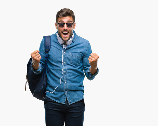 Young handsome tourist man wearing headphones and backpack over isolated background celebrating surprised and amazed for success with arms raised and open eyes. Winner concept. - Fotografie, Obrázek