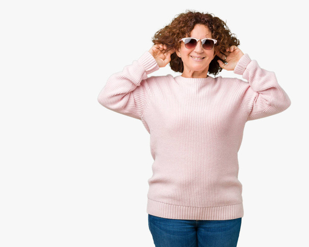Красивая пожилая женщина среднего возраста в розовом свитере и солнцезащитных очках на изолированном фоне, улыбающаяся, дергающая за уши пальцами, смешной жест. Задача прослушивания
 - Фото, изображение