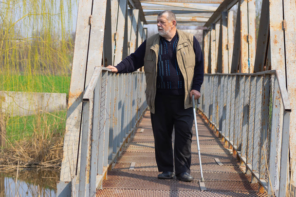 Senior man with walking stick standing on an metal bridge over small river - Photo, Image