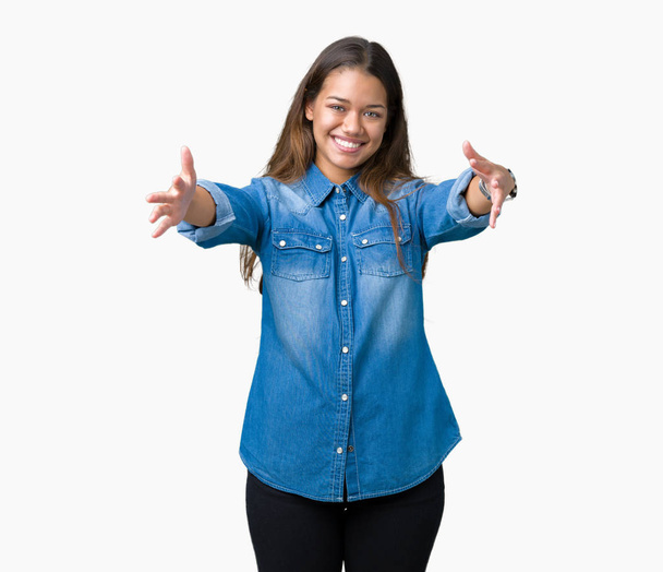 Giovane bella donna bruna che indossa camicia di jeans blu su sfondo isolato guardando la fotocamera sorridente a braccia aperte per l'abbraccio. Espressione allegra che abbraccia la felicità
. - Foto, immagini