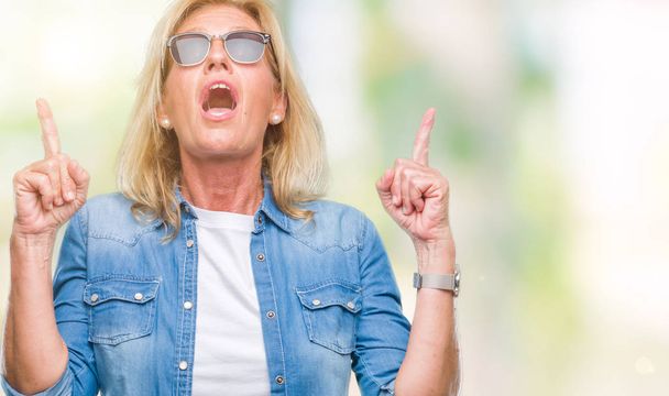 Blonde Frau mittleren Alters mit Sonnenbrille über isoliertem Hintergrund staunte und überraschte Blicke nach oben und zeigte mit erhobenen Fingern und Armen. - Foto, Bild