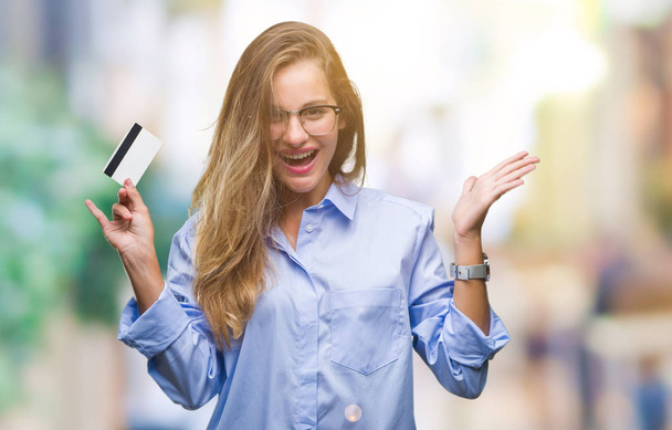 Young beautiful blonde woman holding credit card over isolated background very happy and excited, winner expression celebrating victory screaming with big smile and raised hands - Photo, Image