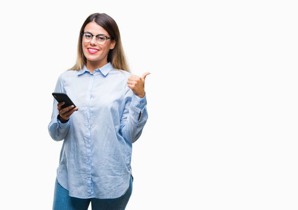 junge schöne Geschäftsfrau SMS mit Smartphone über isolierten Hintergrund zeigt und zeigt mit dem Daumen zur Seite mit glücklichem Gesicht lächelnd - Foto, Bild