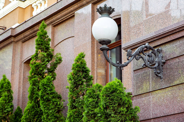 Forged iron street lamp with a white glass ceiling mounted on the marble wall of the facade of the building. - Zdjęcie, obraz