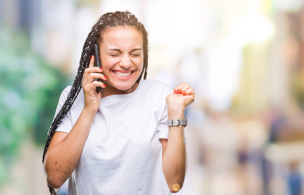 Young gevlochten haar Afrikaanse Amerikaans meisje weergegeven: bellen via smartphone over geïsoleerde achtergrond schreeuwen trots en vieren overwinning en succes erg enthousiast, juichende van emotie - Foto, afbeelding