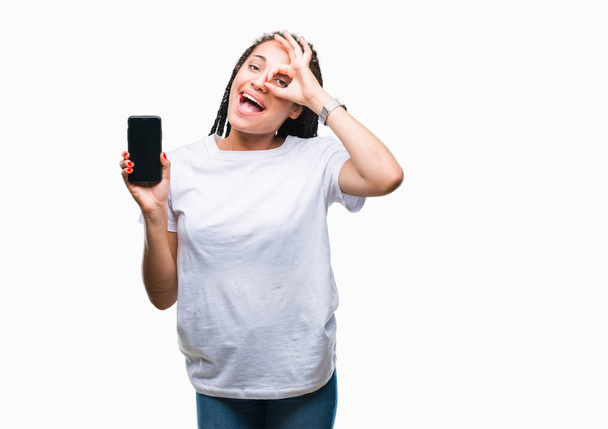 Młody pleciony włosy african american girl pokazano ekran Smartphone na białym tle szczęśliwy twarz uśmiechający się robi ok znak ręką na oko patrząc przez palce - Zdjęcie, obraz