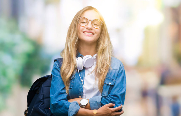 Jonge mooie blonde student vrouw hoofdtelefoons en bril dragen over geïsoleerd achtergrond blij gezicht lachend met gekruiste armen kijken naar de camera. Positief persoon. - Foto, afbeelding