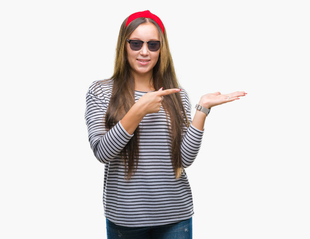 Young beautiful caucasian woman wearing sunglasses over isolated background amazed and smiling to the camera while presenting with hand and pointing with finger. - Photo, image