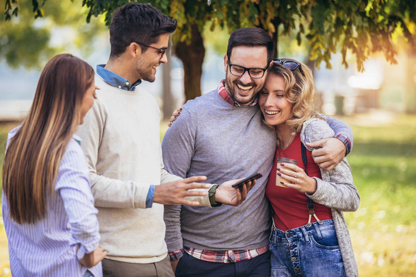 vier fröhlich lächelnde junge Freunde, die draußen im Park mit einem digitalen Tablet spazieren gehen - Foto, Bild