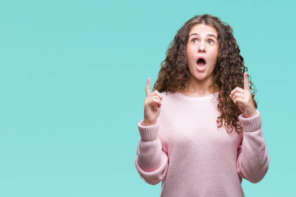 Beautiful brunette curly hair young girl wearing pink winter sweater over isolated background amazed and surprised looking up and pointing with fingers and raised arms. - Photo, Image