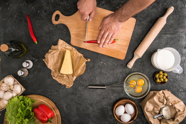 cropped view of man cutting chilli pepper on cutting board with pizza ingredients on grey background  - 写真・画像