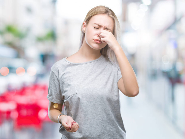 Young blonde woman over isolated background smelling something stinky and disgusting, intolerable smell, holding breath with fingers on nose. Bad smells concept. - Photo, Image