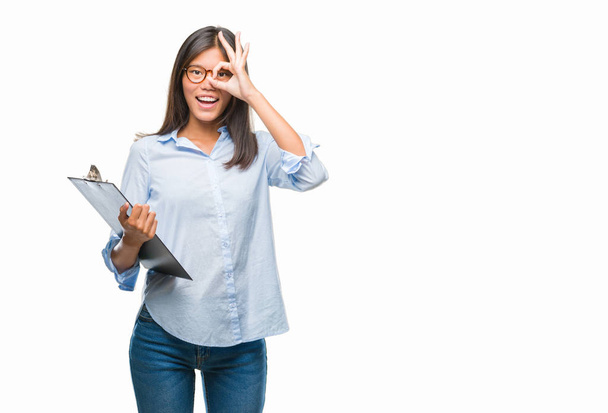 Young asian business woman over isolated background holding clipboard with happy face smiling doing ok sign with hand on eye looking through fingers - Фото, изображение