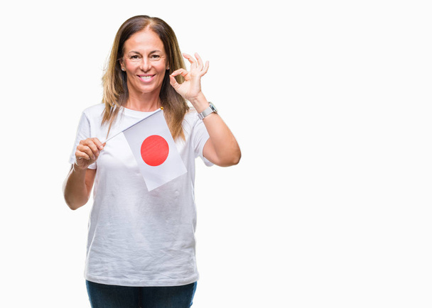 Mulher hispânica de meia idade segurando bandeira do Japão sobre fundo isolado fazendo sinal ok com os dedos, excelente símbolo
 - Foto, Imagem