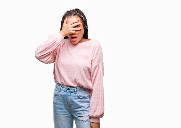Молодая афроамериканка с плетеными волосами в свитере на изолированном заднем плане, заглядывая рукой в лицо и глаза, смотрит сквозь пальцы со смущенным выражением лица
. - Фото, изображение