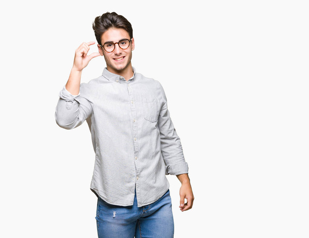 Młody przystojny mężczyzna na białym tle uśmiech i pewność gestem ręką robi znak rozmiar palcami patrząc i aparat w okularach. Pojęcie środka. - Zdjęcie, obraz