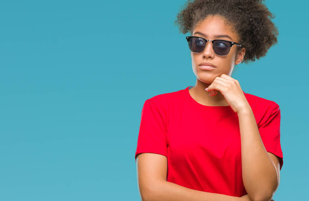 Fiatal afro-amerikai nő napszemüveget visel, mint elszigetelt háttér kézzel a chin gondolkodás kérdés, töprengő kifejezés. Mosolygós, figyelmes arccal. Kétség koncepció. - Fotó, kép